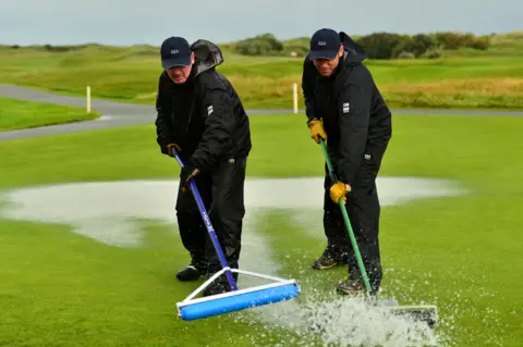 R&A via Getty Images Two people sweep water from a golf green