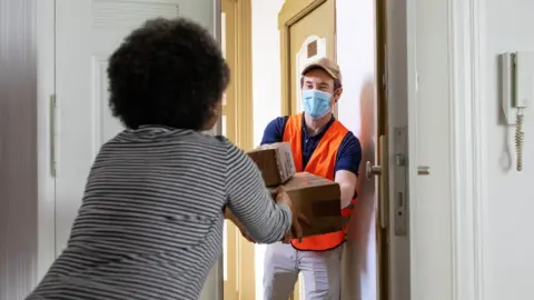 Getty Images Staged photo of a delivery driver handing over a parcel (with PPE and social distancing)