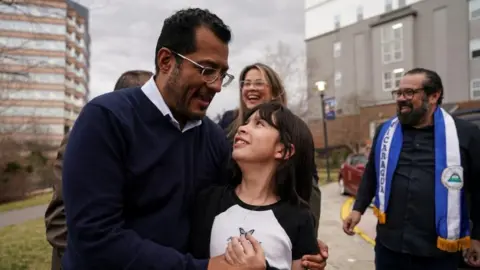 A man wearing glasses hugs his daughter with people looking on