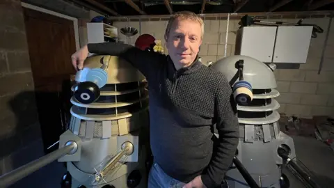 John Chapman wearing a grey jumper and leaning an arm over one of the recovered daleks while standing between them.