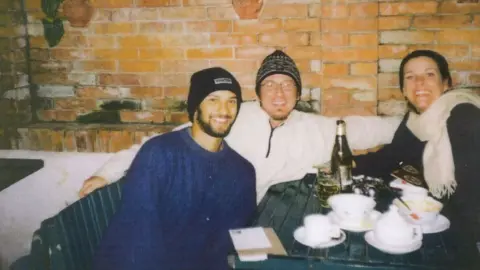 Anna Crozier Last photo of Alex in Nepal wearing a black hat and blue jumper with a beard. He is seated at a table with two others