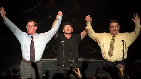 Pacemaker David Trimble, John Hume and U2's Bono raise their hands together at Belfast's Waterfront Hall