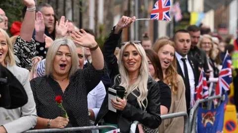 PA Media A crowd of people - some waving, holding flags and cheering - gathered outside Hillsborough Castle ahead of the visit by the King and Camilla