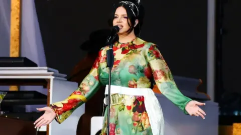 Getty Images Lana Del Rey singing on stage, she is wearing a green floaty dress and her arms are open. She is wearing her black hair up.