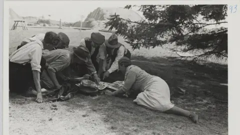 Barbara Wagstaff A.R.P.S/Trustees of the British M Mercie Lack showing members of the excavation team a selection of contact prints, with the excavation in the background