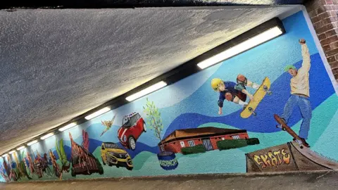 Sarah Harris An underpass with a mural on one side, with a background in different shades of blue and skateboarders, cars and dragons painted