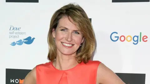 Getty Images A woman with shoulder length blonde hair smiles widely for cameras as she stands in front of a red carpet hoarding bearing the logos of Google and Dove soap. She's wearing a red-orange sleeveless dress and sparkly earrings.