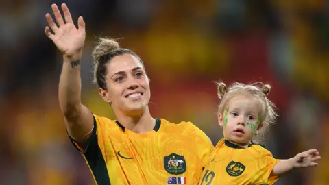 Getty Images Katrina Gorry carries two-year-old daughter Harper, while wearing matching football kits, as they wave to the crowd