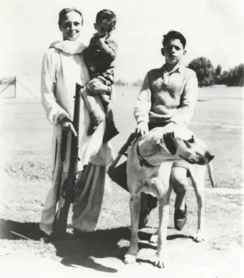 Freda with a rifle when she was living in Kashmir, probably 1948 - she is holding hr son, Kabir, while her older son, Ranga, is sitting on the family's pet dog.