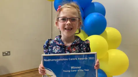 Martin Heath/BBC Young girl with glasses and hair in pony tails holds an award certificate