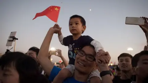 Getty Images A man carries his son on his shoulders to see the lower of the flag on Tiananmen square in Beijing on September 28, 2017.