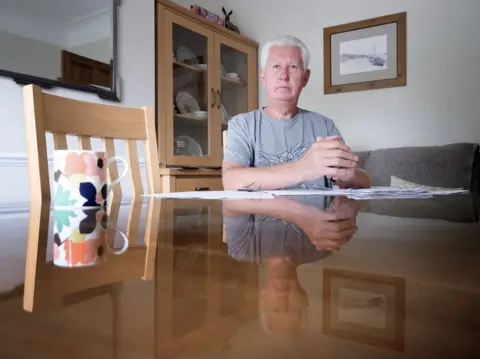 BBC Ian Ross shown with a reflection of himself in the table. He looks pensive and is surrounded by paperwork