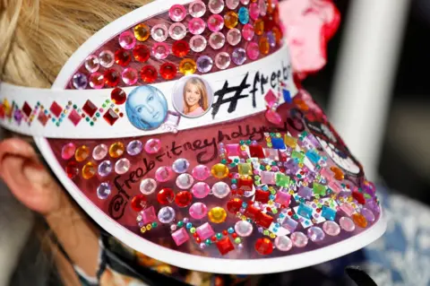 Mario Anzuoni / Reuters A supporter of pop star Britney Spears wears a visor with buttons of the singer during a gathering on the day of a conservatorship case hearing at Stanley Mosk Courthouse in Los Angeles, California, US