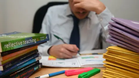PA Media A male teacher leaning his head on his hand whilst surrounded by books