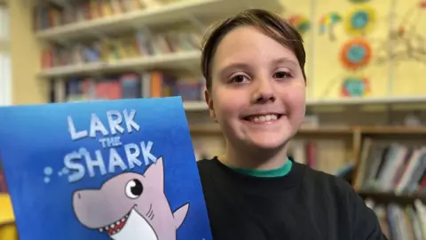 Henry Newman posing with his picture book Lark The Shark.