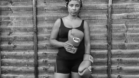 A woman poses against a wooden fence holding boxing gloves