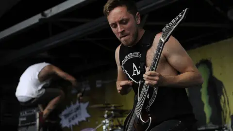 Getty Images Tom Searle stands, playing the guitar on-stage with one knee supporting the instrument. He looks intense as he gears up to strum a chord - he appears to be gently biting his bottom lip as he concentrates. In the background a bandmate can be seen leaping into the air.