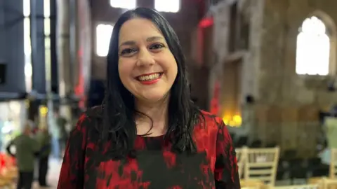 Dr Janina Ramirez in Gloucester Cathedral smiling while wearing a red and black top