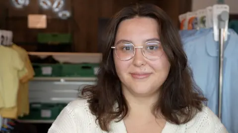 Jamie Niblock/BBC Elisha looking down the camera, standing in the uniform exchange shop.
