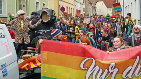 A crowd of people in a huge variety of bright colours and rainbow attire gather in the small street of Ostritz, waving placards and bearing banners