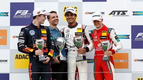 James Gasperotti Enaam Ahmed is stood on a podium with three other racing drivers. He is standing second from the right wearing a white Mercedes racing driving overalls with a white cap. He is smiling and holding a silver trophy in his left hand. Stood next to him are three white racing drivers who are also all wearing racing overalls and holding silver trophies and smiling. Behind them is a sponsors board with various racing sponsors on it.