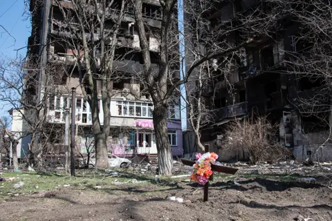 Getty Images 2022/03/26: A freshly dug grave next to two bombed out residential buildings in the besieged city of Mariupol.