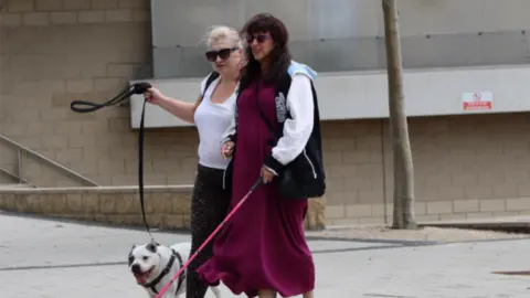 Steve Keenan Photograph of two women walking arm in arm along the pavement. The woman on the left is blonde and holds the lead for a white dog and the woman on the right has brown hair, wears a magenta dress and is holding a pink cane. They both wear sunglasses.