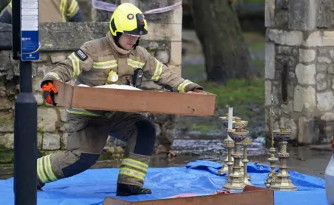 PA Media Image showing a firefighter kneeling down with a drawer of objects, in front of candlesticks laid out on blue tarp.