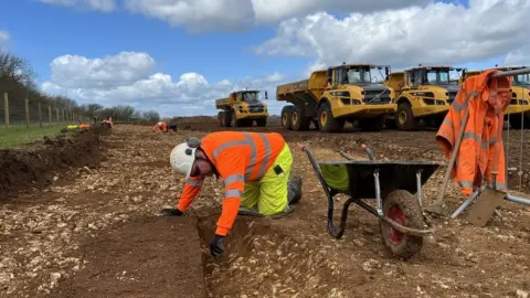 BBC A man digs in the ground with machinery behind him