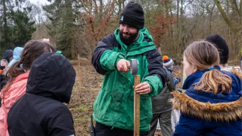 Forestry England A session leader shows the pupils how to put a stake in the ground so the young trees can climb it.
