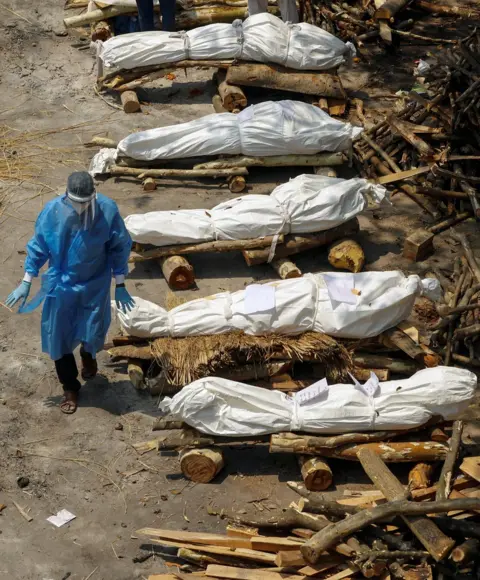 Adnan Abidi / Reuters A health worker wearing personal protective equipment (PPE) walks past a row of wrapped bodies of Covid victims lying on prepared funeral pyres in New Delhi, India