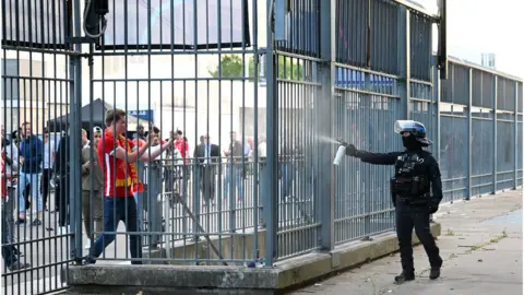 Getty Images Police spray tear gas at Liverpool fans outside the stadium as fans struggle to enter prior to the UEFA Champions League final match between Liverpool FC and Real Madrid at Stade de France on May 28, 2022 in Paris, France. (Photo by Matthias Hangst/Getty Images)
