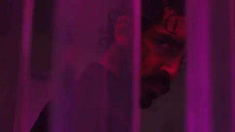 Universal Dev Patel as 'Kid' in Monkey Man. Dev, a 33-year-old British Asian Man, looks at the camera with a sinister expression over his shoulder. A few locks of his curly dark hair flop over his face and he's wearing a dark blazer, facing away from the camera. He's lit by purple lighting and is slightly obscured by thin purple columns