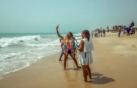 Grace Ekpu A woman on a beach taking a selfie