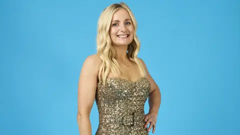 Matt Frost A woman in a golden, shoulderless, sparkly dress stands in front of a light blue background, smiling widely with one hand resting on her hip.