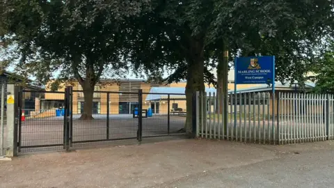 BBC Image of Marling School in Stroud. There is a closed metal gate in front of the building and lots of trees in the area. The school signpost is blue, and reads "Marling School West Campus"