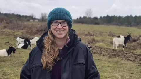 Kayleigh Poacher/BBC Laura Davey stood in a field surrounded by goats