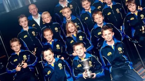 Maddy Cusack foundation West Hallam juniors football team, a group of young children sit in blue tracksuits. One of the boys is holding a trophy. Maddy is the only girl in the photo.