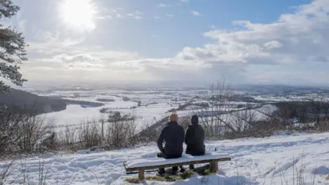 Getty Images A couple sat on a bench overlooking the snow-covered fields of Thirsk at Sutton Bank National Park Centre in the North York Moors