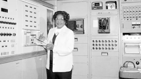 NASA Mary Jackson holds a clipboard standing by a large computer