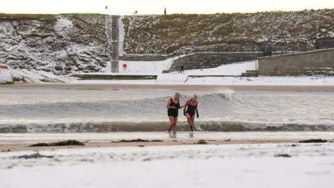 PA Media Cold-water swimmers walk across a snow-covered beach at Cullercoats Bay on the North-East coast