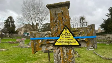 BBC A cross headstone with a yellow warning taped onto it with blue tape. The warning reads: "Warning! This memorial is unsafe. Do not tamper with temporary support. Please contact Cemetery Office."