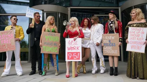 PA Media A line of eight tribute artists outside the Meta office, with Dolly Parton centre holding a megaphone