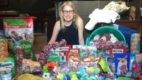 A young girl sits in front of lots of dog food, dog biscuits, dog toys, etc.