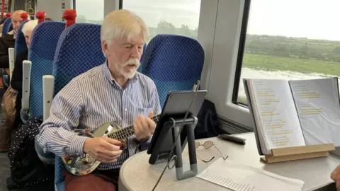 BBC A man plays a ukulele while sitting on a train