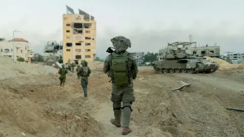 Reuters Israeli soldiers on patrol in the Gaza Strip. They are seen walking in single file across muddy ground towards a number of damaged buildings.