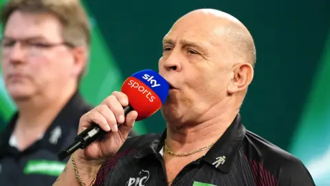 PA Media A man wearing gold chains holds up and speaks into a blue and red Sky Sports microphone