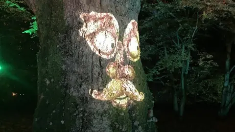 BBC An older face with a moustache lit up onto a large tree trunk