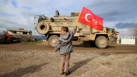 Getty Images A photo taken from Turkey's Hatay province shows children greets soldiers with Turkish flags during Turkish military convoy consisting of approximately 300 armoured personnel carriers are on the way towards observation points in Syria's Idlib, on February 08, 2020 in Hatay, Turkey