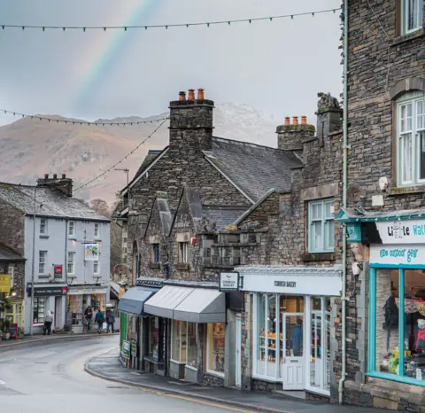 Liam Norman A view of shops in old stone buildings on a street with hills and a rainbow in the distance beyond